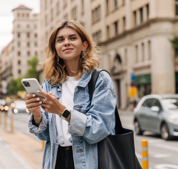 Picture of pretty young woman staying on the street holding phone in hands. Stylish blonde looking away and smiling in city. Use technology concept